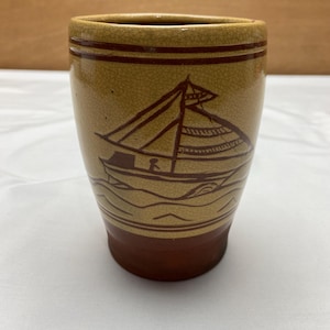 May include: A brown ceramic vase with a beige glaze and a brown sailboat design. The vase has a textured surface and a wide mouth.