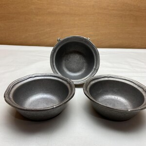 May include: Three small, antique-style pewter bowls. Two are oval-shaped, and one is round with a small handle. All have a decorative scalloped edge. The bowls are arranged on a white surface with a light brown background.