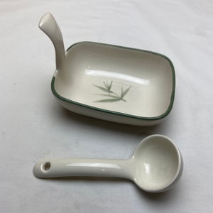 May include: A white ceramic serving dish with a green rim and a green leaf design. The dish has a handle on one side. A white ceramic ladle is also included.