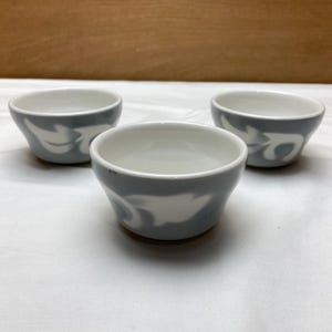 May include: Three small, gray and white ceramic bowls. Each bowl has a gray exterior with a white, abstract design. The interior of each bowl is white. The bowls are arranged on a white surface.