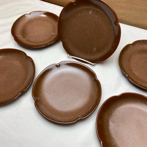 May include: Six brown ceramic plates with scalloped edges. The plates are arranged in a semi-circle on a white surface.