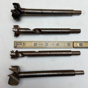 May include: Four dark grey metal Forstner drill bits of varying diameters arranged horizontally. A yellow wooden folding ruler is positioned between the second and third bits, showing a measurement scale in inches from one to six inches.