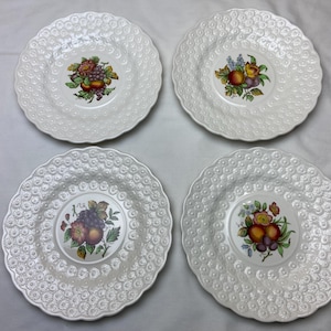 May include: Set of four white ceramic plates with scalloped edges and embossed designs. Each plate features a different floral and fruit motif in the center.