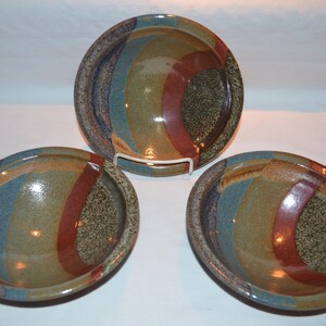 May include: Three handmade ceramic bowls with a colorful design. The bowls feature a mix of colors, including blue, brown, and red, arranged in curved patterns. The bowls are displayed on a white surface.