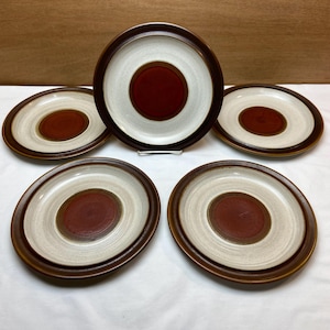 May include: A set of five ceramic plates. Each plate features a dark brown rim, a light beige outer ring, and a deep red center. The plates are arranged on a white surface, with a brown background.