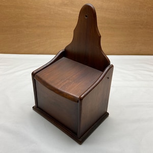 May include: A dark brown wooden wall-mounted salt box with a decorative top. The box has a rectangular base and a hinged lid. The wood grain is visible, and the box appears to be vintage.