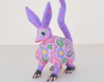 Könnte beinhalten: Handgeschnitzte, farbenfroh bemalte Holzskulptur eines Alebrije, die ein kaninchenartiges Wesen darstellt. Die Skulptur hat einen rosa Körper mit Blumen- und Punktmustern, lila Ohren und Schwanz sowie schwarze Füße. Das Wesen hat ein Schlangendesign auf dem Rücken.