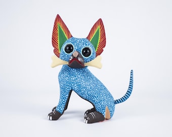 May include: A blue and white spotted dog figurine with large, colorful ears and a bone in its mouth. The folk art sculpture has black eyes, a black nose, and brown feet. The dog's tail is striped blue and white.