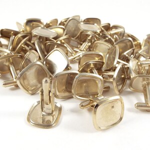 May include: A pile of gold-toned cufflinks with square-shaped glass inserts. The cufflinks are arranged in a random pattern.