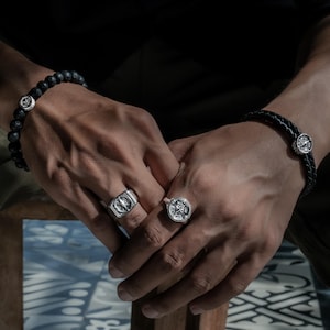 Puede incluir: Un conjunto de joyería compuesto por dos anillos de plata con motivos de brújula circulares, una pulsera de cuentas de lava negra con un dije de brújula de plata y una pulsera de cuero trenzado negro con un dije de brújula.