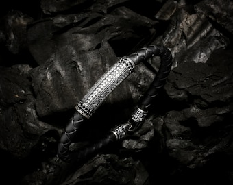 Op de afbeelding: Een gevlochten zwarte leren armband met een centrale zilverkleurige metalen cilinder met ingewikkelde gegraveerde patronen en kleine zwarte steentjes, afgewerkt met een bijpassend metalen sluitingsmechanisme.