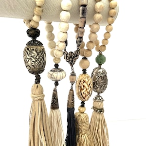 May include: Five beaded necklaces with tassels. Each necklace features a different pendant, including a carved bone pendant, a silver pendant, and a green stone pendant. The necklaces are made of white and beige beads.