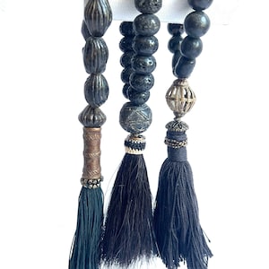 May include: Close-up of three beaded tassels. The beads are dark grey and black, with various textures and shapes. Each tassel has a different design, including metal accents and dark-coloured thread. The tassels hang against a white background.