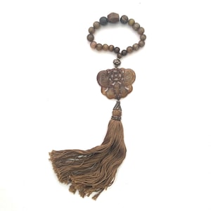以下が含まれることがあります： 彫刻された蝶のチャームとビーズのブレスレットが付いた茶色のタッセル。蝶のチャームは細部までこだわったデザインで、薄い茶色の素材で作られています。ブレスレットは茶色のビーズで作られており、端に茶色のタッセルが付いています。