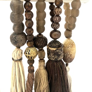 Pode incluir: Um conjunto de cinco colares de contas com borlas. Os colares são feitos de contas marrons, bege e brancas. As borlas são feitas de fio marrom, bege e branco.