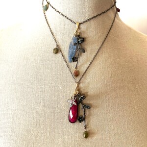 Puede incluir: Dos collares en capas con delicados acentos florales. El collar superior presenta un colgante de piedra azul, mientras que el collar inferior presenta un colgante de piedra roja.