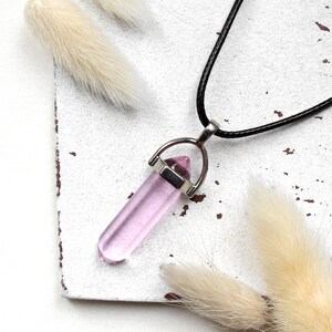 Könnte beinhalten: Eine Halskette mit einem rosa Kristall-Anhänger mit silberner Fassung an einem schwarzen Band. Die Halskette liegt auf einer weißen Holzoberfläche mit getrockneten weißen Blumen.