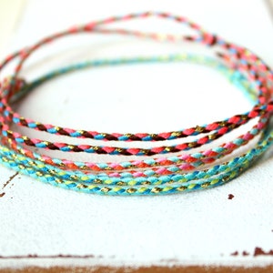 Könnte beinhalten: Ein Set aus fünf geflochtenen Schnüren in Türkis, Rosa, Braun und Gold. Die Schnüre sind miteinander verwoben und haben einen dezenten Schimmer.