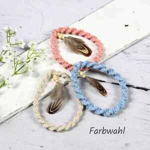 Può includere: Tre braccialetti intrecciati in corda con perline di legno e piume. I braccialetti sono rosa, blu e naturali. Il testo "Farbwahl" è visibile nell'immagine.