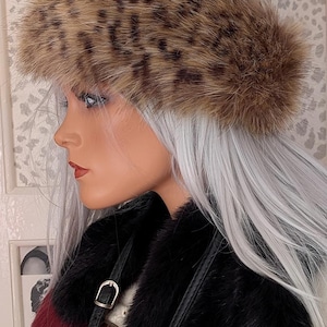 Può includere: Fascia per capelli in finta pelliccia con motivo leopardato. La fascia è marrone e beige con macchie nere. Il manichino indossa un cappotto di pelliccia nero con una striscia rossa. Il manichino ha i capelli argentati e rossetto rosso.