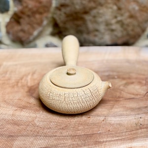 Puede incluir: Una pequeña tetera de cerámica beige con asa y boquilla. La tapa es redonda con un pequeño pomo. La tetera tiene una superficie texturizada y agrietada. La tetera está sobre una superficie de madera.