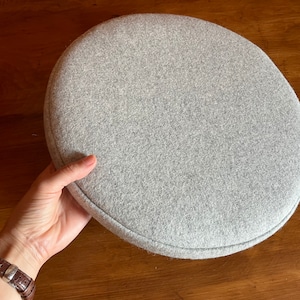 May include: A gray round cushion with a stitched edge. The cushion is made of a soft, fuzzy fabric.
