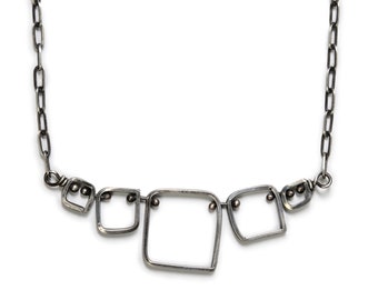 Puede incluir: Un collar de plata con un diseño geométrico que presenta cinco cuadrados conectados. Los cuadrados están abiertos y tienen pequeñas cuentas de plata en las esquinas.
