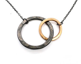 May include: A necklace featuring two interlocking rings. One ring is a dark gray color, and the other is a gold color. The rings are connected by a dark chain. The necklace is displayed on a white background.