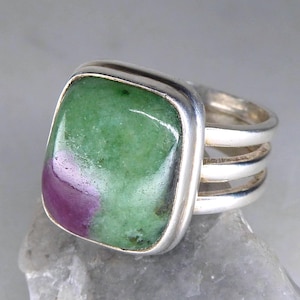 May include: A silver ring with a large, square gemstone. The gemstone is a mix of green and purple. The ring has a triple-band design. The ring is displayed on a grey stone surface.