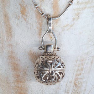 May include: A silver-toned spherical pendant featuring an intricate openwork filigree pattern, suspended from a matching silver-toned chain. The round locket-style charm has a hinged bail and a secure latch mechanism, showcasing detailed metalwork.
