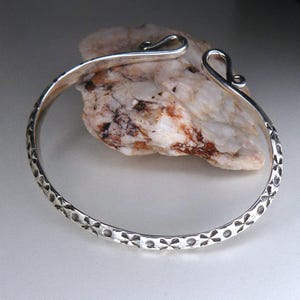 May include: A silver bangle bracelet with a hammered finish and a floral design. The bracelet has a simple hook closure.