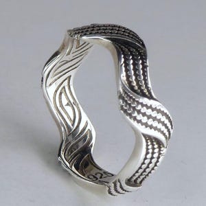 May include: A silver ring with a wavy, undulating band featuring a textured pattern of small raised dots on one side and smooth, carved linear grooves on the other. The interior of the band is clearly stamped with the hallmark 925.