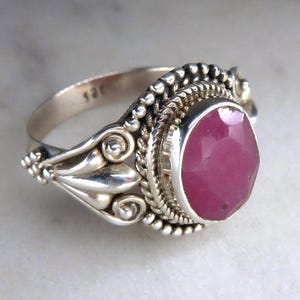 May include: A silver ring featuring a faceted oval ruby gemstone. The ring has an intricate design with beaded and scrollwork details. The ruby is a deep pinkish-red colour, and the ring is set on a light-coloured surface.