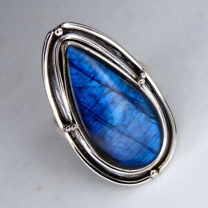 May include: A teardrop-shaped labradorite gemstone ring set in a polished silver frame. The stone displays a vibrant blue iridescence with visible internal fractures. The metal setting features a layered design with small silver beads at the edges.