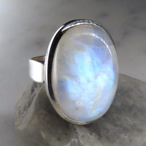 May include: A silver ring featuring a large, oval-shaped moonstone cabochon. The stone displays a translucent white appearance with iridescent blue flashes across its surface. The metal band is smooth, polished, and encircles the stone setting.