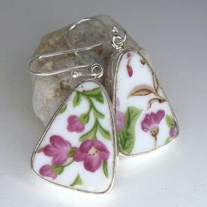 May include: A pair of silver earrings with triangular shaped porcelain pendants. The pendants feature a floral pattern with pink flowers and green leaves on a white background.