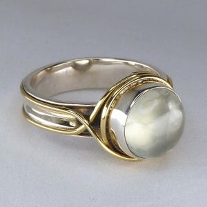 May include: A ring featuring a round, pale green cabochon gemstone set in a polished silver bezel. The band is constructed from silver with decorative gold-toned wire wrapping that crosses over the sides and encircles the central stone setting.