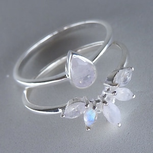 May include: A silver ring featuring a pear-shaped moonstone and a cluster of smaller moonstones. The ring has a double-band design, with one band curving around the finger and the other with gemstones.