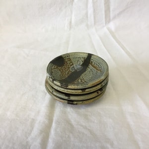 May include: A stack of five small, round ceramic plates. The plates have a speckled, earthy color palette with brown, black, and tan tones. Each plate features a unique, textured design.