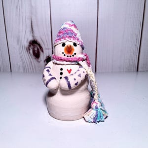 May include: A small white snowman figurine with a pink and purple crocheted hat and scarf. The snowman has a black button nose and a red heart on its chest.