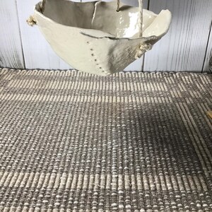 May include: A handmade, off-white ceramic hanging planter with a textured surface. The planter is suspended by three natural fiber ropes. The design features a unique, asymmetrical shape and is suitable for small plants or succulents.