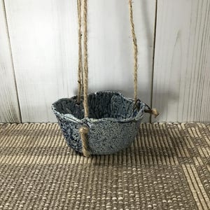 May include: A hanging planter with a blue and white floral design. The ceramic pot is suspended by natural rope. The planter is empty and ready for a small plant. The background is a light wood panel and a woven rug.