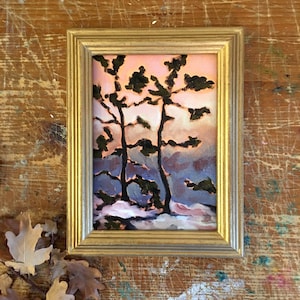May include: A framed landscape painting depicting two trees silhouetted against a sunset. The artwork uses pinks, blues, and greens, with a gold-coloured frame. The painting evokes a serene, natural scene, suitable for home decor.