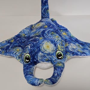 Può includere: Un giocattolo di peluche a forma di manta, con un motivo a vortice blu e giallo che ricorda "La notte stellata" di Van Gogh. Il giocattolo ha grandi occhi espressivi con dettagli verdi e bianchi e un lato inferiore bianco. Le ali sono spiegate.