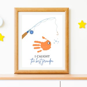May include: Framed art print featuring a handprint fish design with a fishing rod illustration. The text reads "I caught the best grandpa" in blue script. The frame is made of wood.