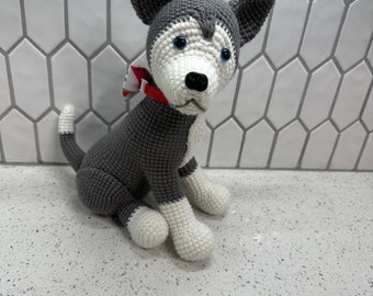 May include: A crocheted husky dog plush toy with gray and white fur, blue eyes, and a red collar with a patterned bow. The toy is sitting on a white countertop.
