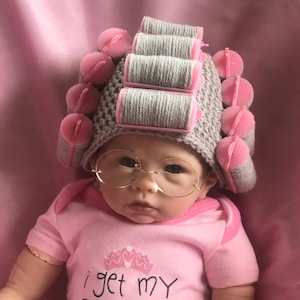 May include: A baby wearing a pink onesie with the text "i get my style from my" and a grey and pink crocheted hat with grey hair rollers. The baby is also wearing round, clear glasses.