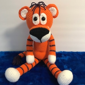 May include: A handmade orange and black striped crochet tiger toy with white accents. The tiger has large black eyes, a black nose, and white paws. The tiger is sitting on a blue surface.