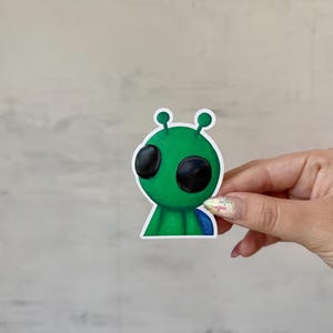 May include: A green alien sticker with large black eyes and two antennae. The alien has a blue cape and is held by a hand. The sticker is a cartoon illustration.