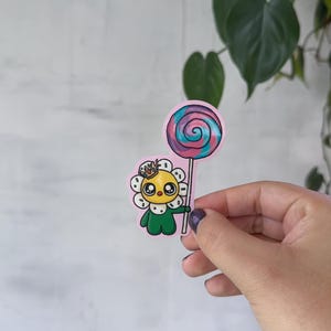 May include: A sticker featuring a cartoon sunflower character wearing a crown and holding a lollipop. The sunflower has a yellow face, white petals, and a green body. The lollipop is swirled with pink, blue, and purple colors.